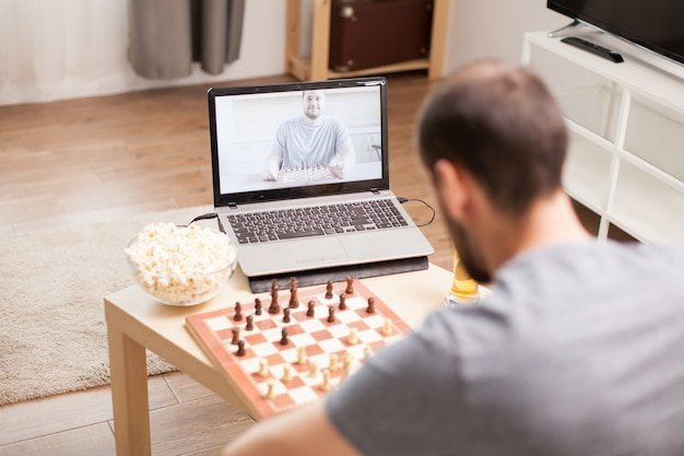 Two men playing a remote chess game over a video call on a laptop, with a physical chessboard, popcorn, and a drink.