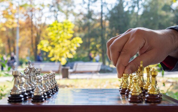 Close-up of a hand moving a gold chess pawn on a board. Silver pieces are opposite, set in a vibrant autumn park.