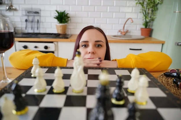 Young woman in yellow sweatshirt intently looks at a chessboard in a bright kitchen, a glass of red wine nearby.
