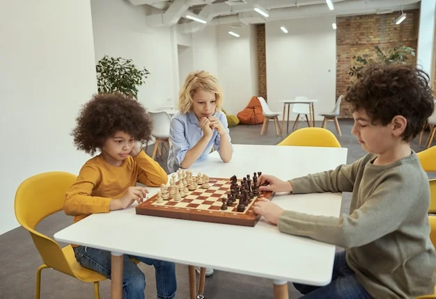 Three young boys intently playing chess at a white table in a bright room, with one boy observing the game.