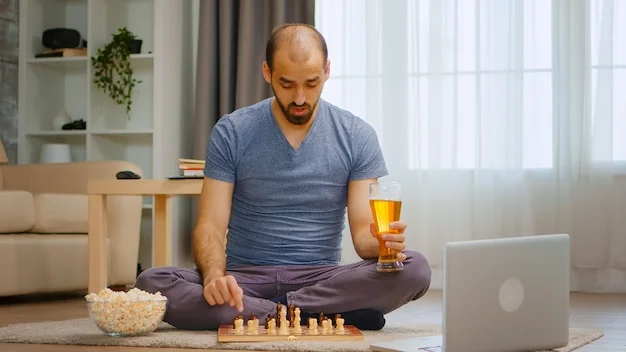 Focused man playing chess on the floor, holding a beer. A bowl of popcorn and a laptop are beside him in a home setting.