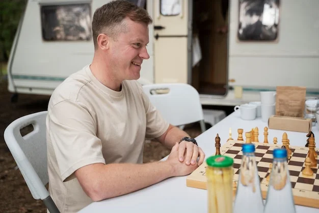 Smiling man enjoying a game of chess at a table outdoors, with a white camper van in the background. Leisurely camping scene.